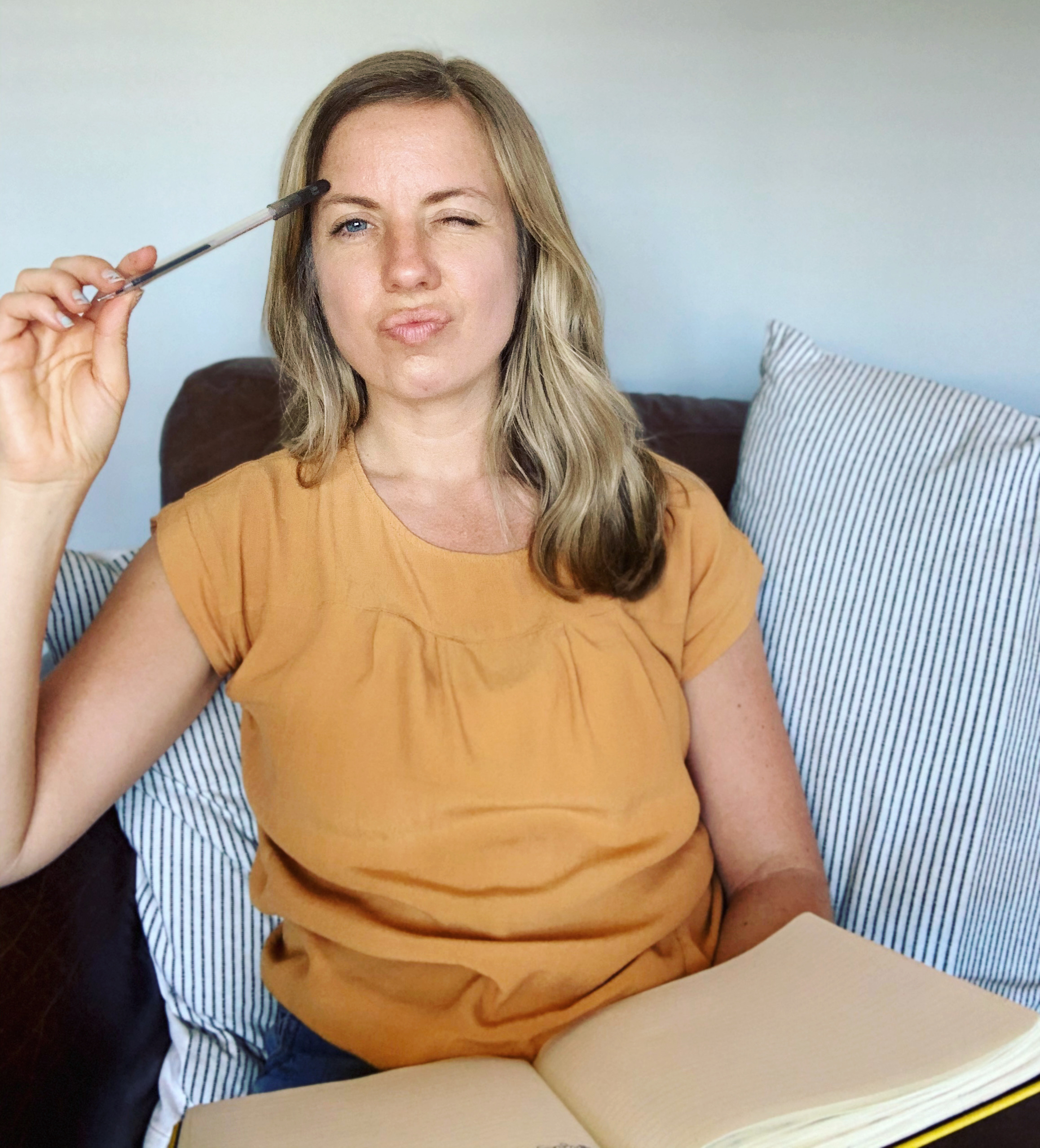 A woman is sat on a sofa, with a notebook in hand. She is resting her closed pen on her temple, looking very confused. She has blonde wavy hair and a short sleeve orange top. She looks overwhelmed with the amount of pricing options there are!
